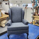 Wingback chair reupholstery featuring a classic high-back armchair in textured blue-grey fabric with elegant black cabriole legs in a professional workshop