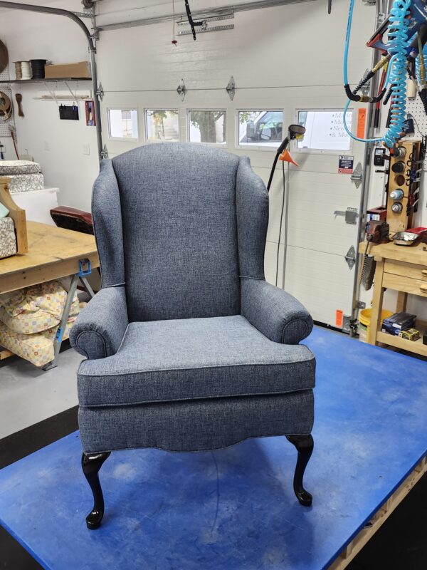 Wingback chair reupholstery featuring a classic high-back armchair in textured blue-grey fabric with elegant black cabriole legs in a professional workshop