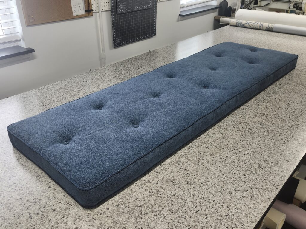 A long, custom-made window seat cushion featuring blue textured fabric and decorative button tufting on a workshop table