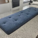 A long, custom-made window seat cushion featuring blue textured fabric and decorative button tufting on a workshop table