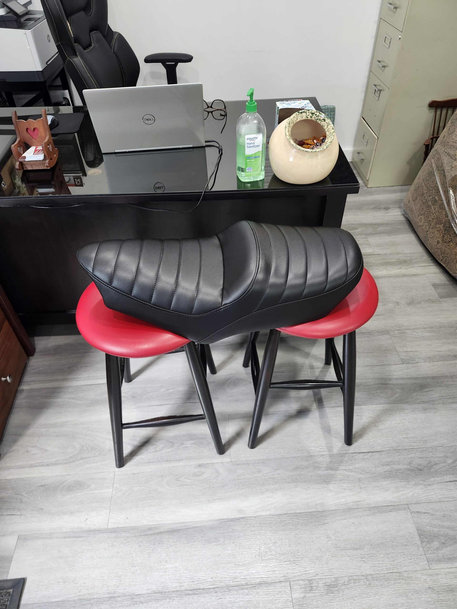 Motorcycle seat upholstery York PA project: long tapered motorcycle saddle recovered in black vinyl with horizontal tuck and roll pleats, resting on red stools at Alex Upholstery Shop