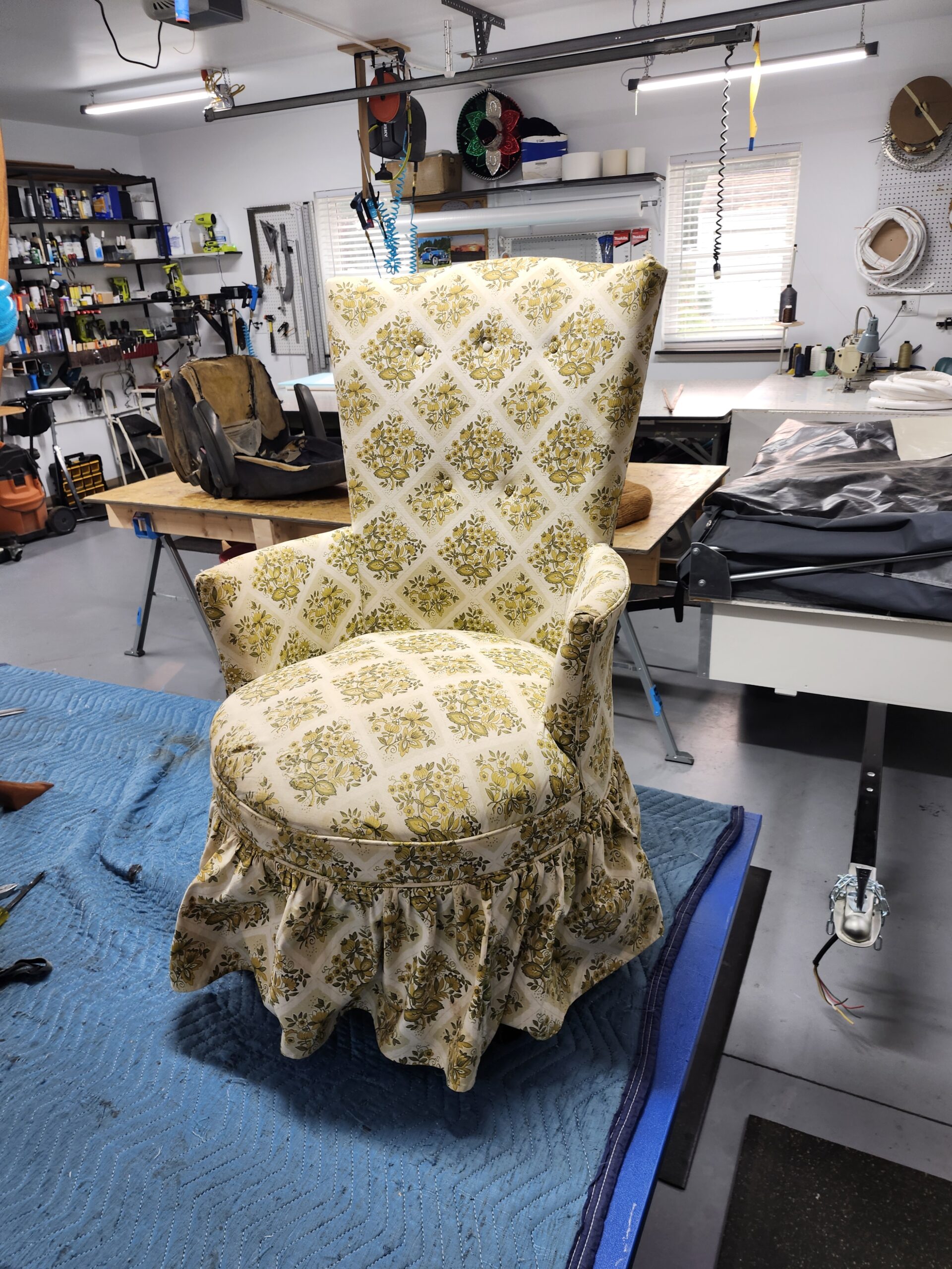 Mid-century round slipper chair before reupholstery in a faded cream, yellow and olive floral diamond lattice print with a five button tufted back, round seat and ruffled skirt to the floor, on the workbench at Alex Upholstery Shop
