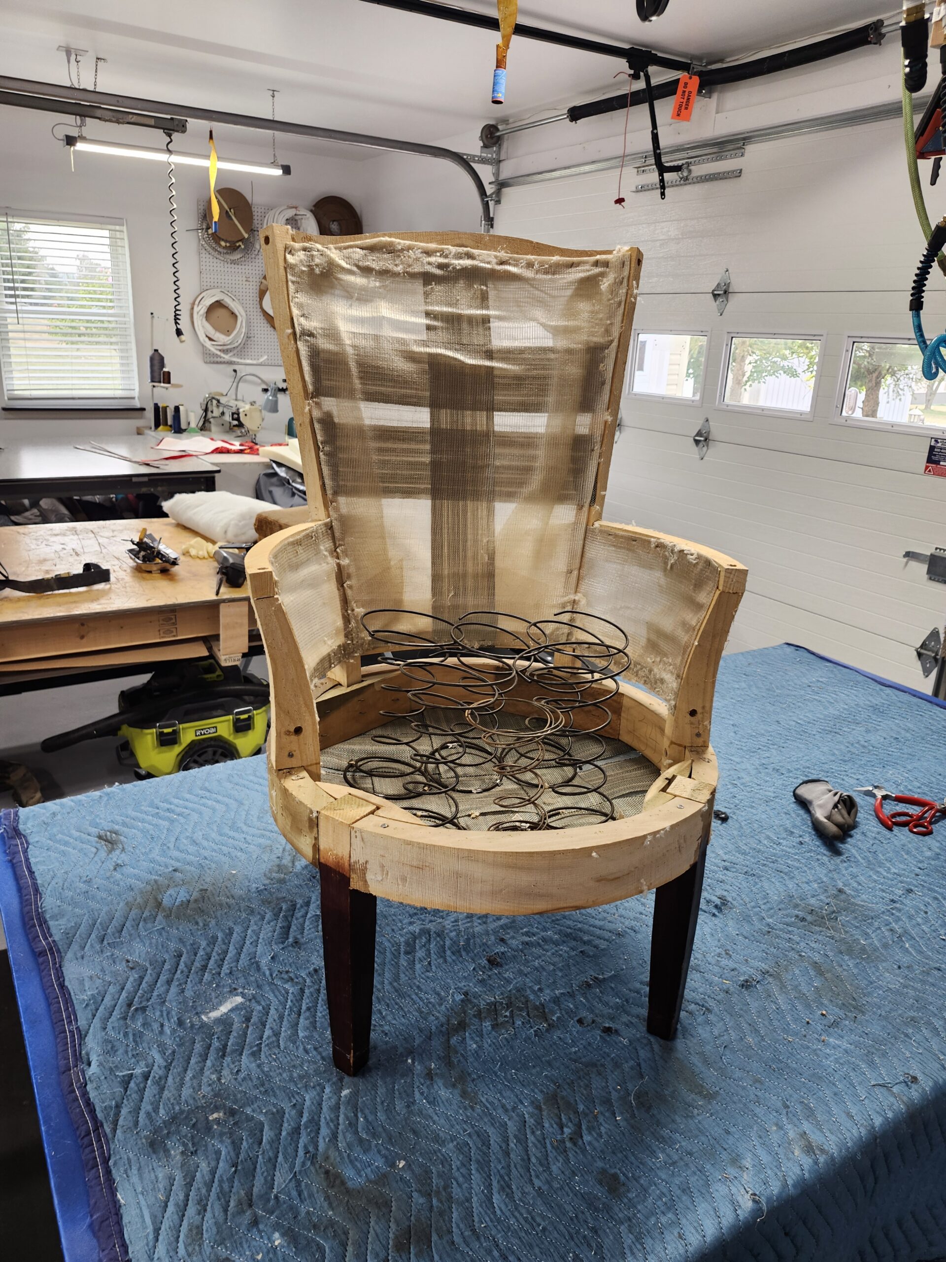 Stripped frame of the round slipper chair on the workbench: a round wooden seat ring with loose coil springs, a tall narrow back frame with jute webbing and burlap still on, low arm frames and dark tapered legs, at Alex Upholstery Shop