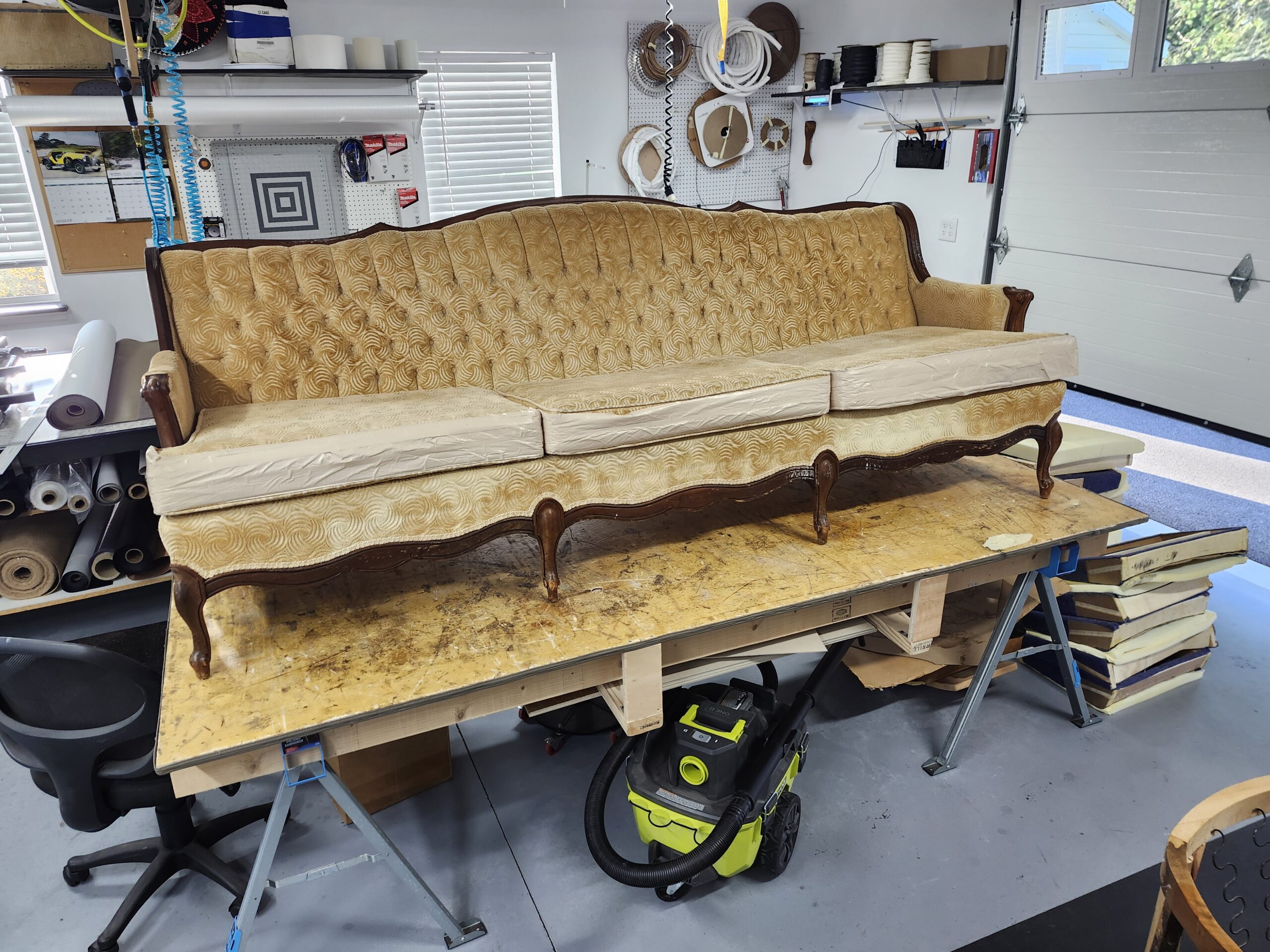 Before: long three cushion French Provincial sofa in worn gold swirl velvet with all three loose seat cushions stripped to bare muslin, diamond tufted back and carved wood trim, on the workbench at Alex Upholstery Shop
