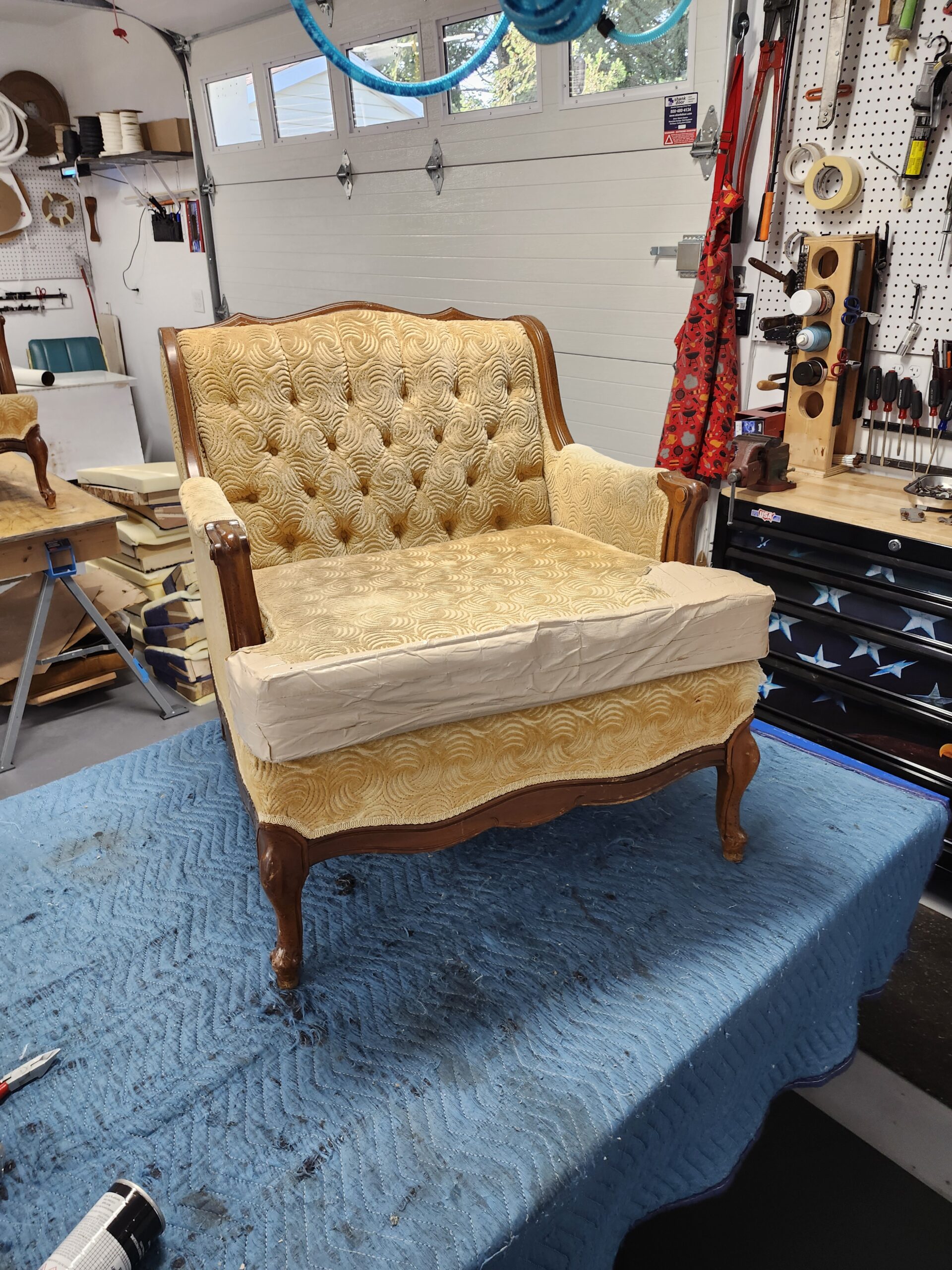 Before: wide French Provincial armchair in gold swirl velvet with diamond button tufted back, scrolled wood arms and cabriole legs, its loose seat cushion stripped to bare muslin, on a blue moving blanket at Alex Upholstery Shop