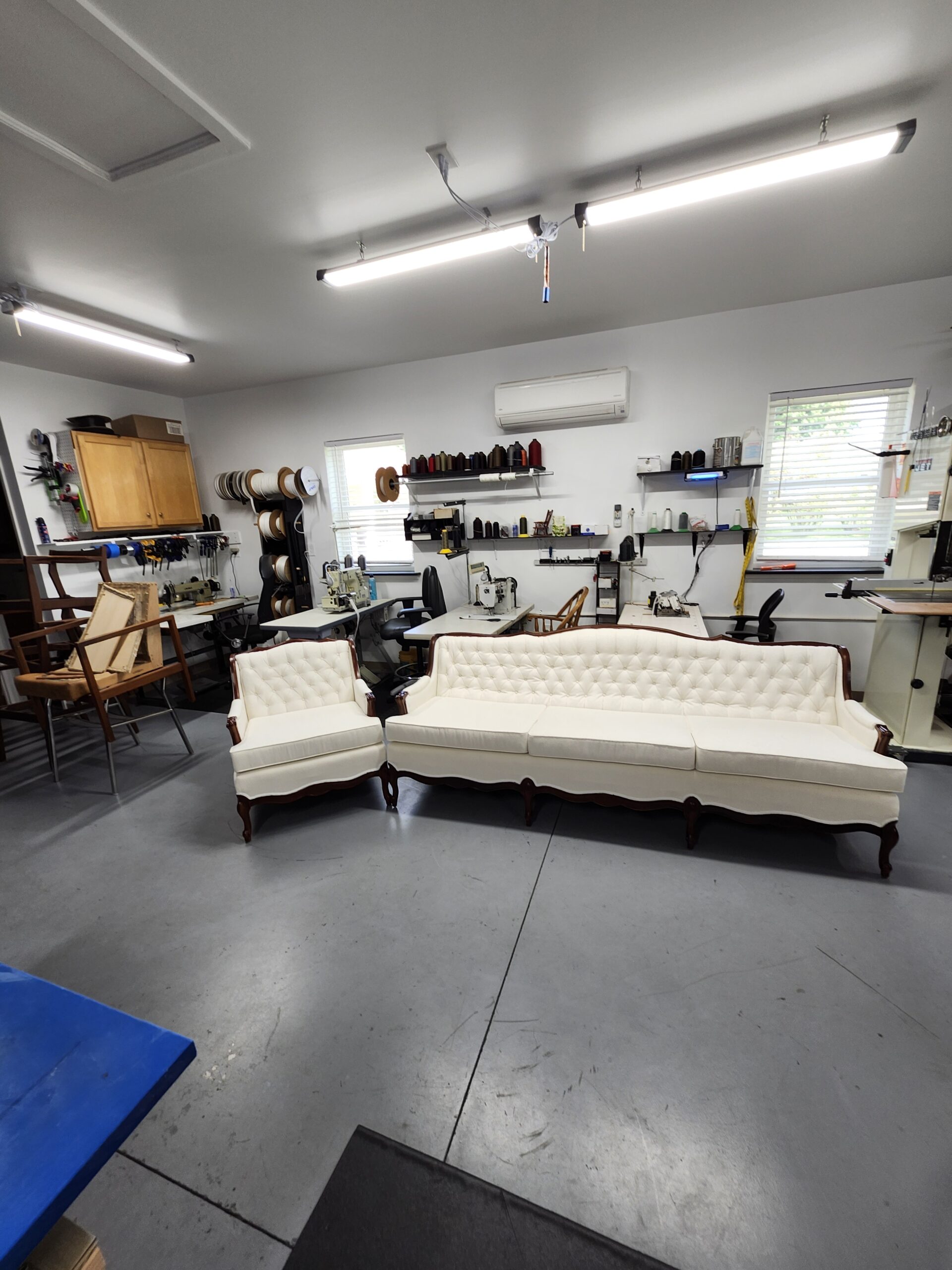 Wide view of the reupholstered ivory French Provincial sofa and chair set standing together in the middle of the Alex Upholstery Shop workroom floor under the shop lights