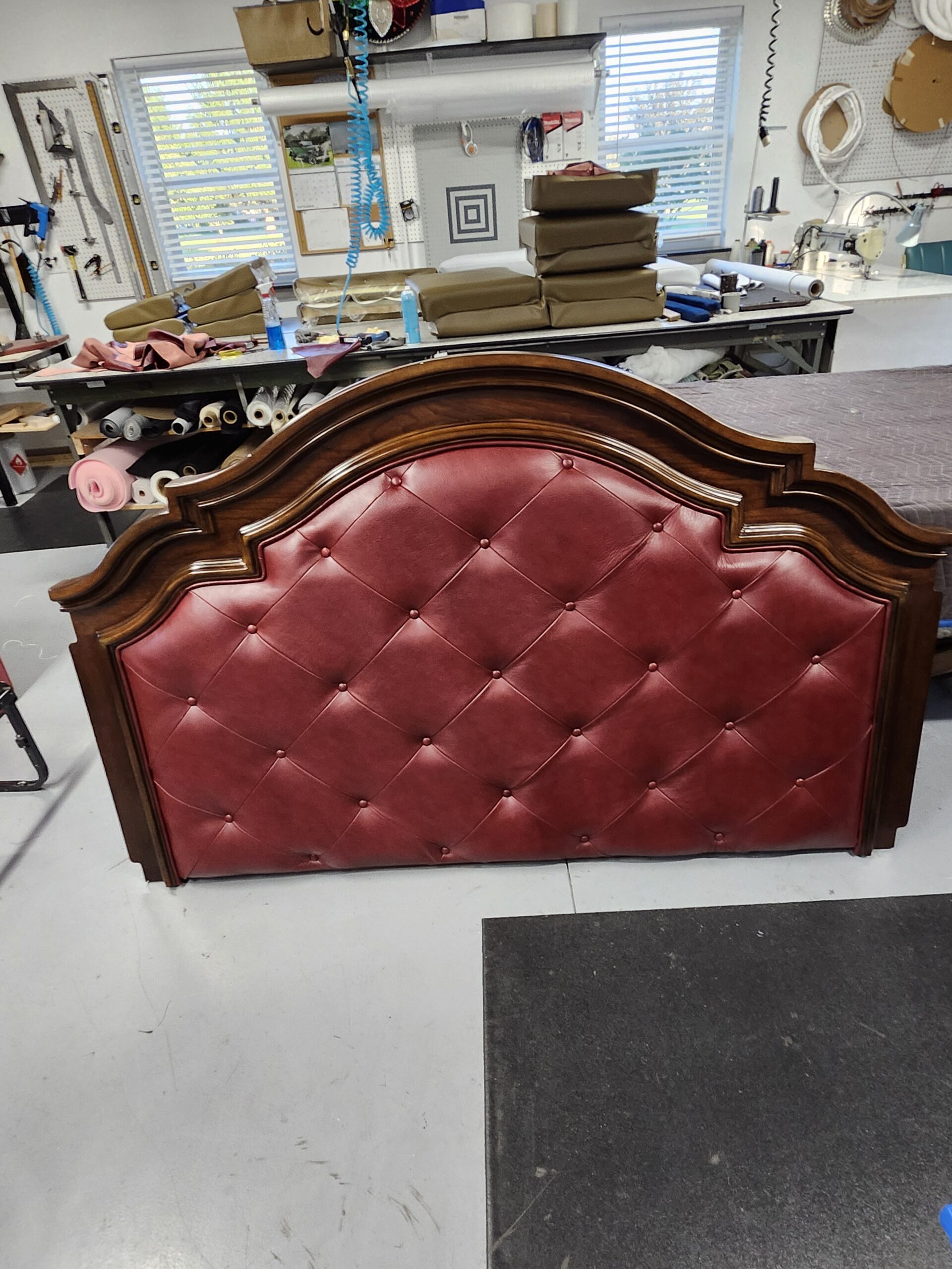 Diamond tufted oxblood leather headboard in an arched wood frame seen from a higher angle on the shop floor, with offcuts of the same hide on the cutting bench behind it at Alex Upholstery Shop