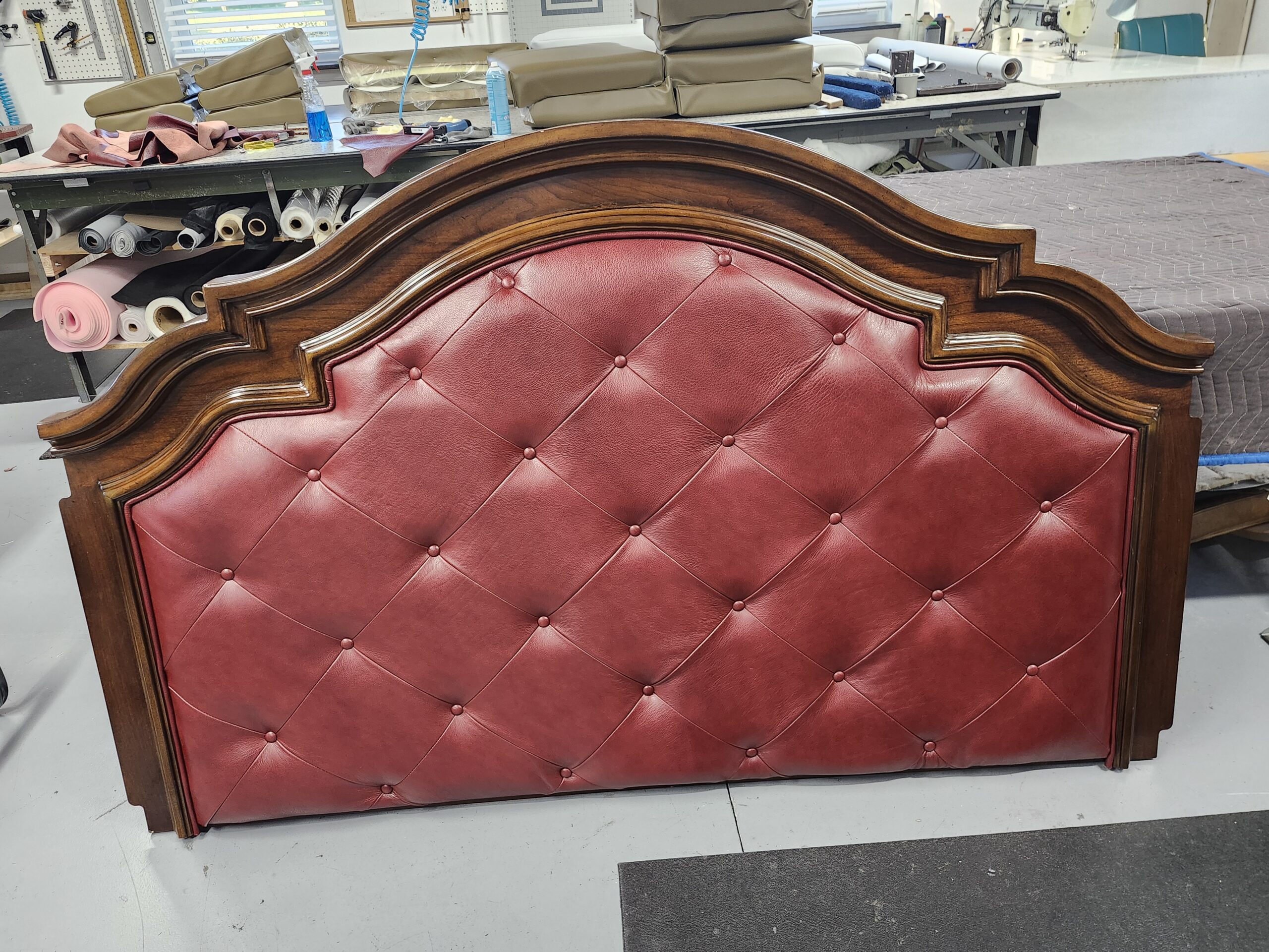Headboard upholstery Mechanicsburg PA project: finished arched wood headboard with a diamond button tufted panel in oxblood pull-up leather, standing on the floor at Alex Upholstery Shop